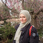 Head shot of Shahdah Mahhouk, a woman wearing a light gray hijab and a black winter coat showing a red bag strap, standing in front of a blossoming tree.