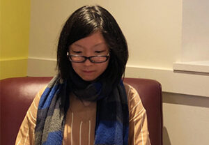 Snow Zhang, sitting, wearing glasses, and looking down at her lap typing, wearing a yellow blouse and blue scarf