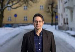 A middle-aged white man with short, mousy brown hair and glasses standing in suit jacket and shirt against a wintery background in a European town.