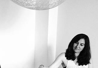 Black-and-white image of Tamar Lando sitting, in the bottom right corner, in a minimalist room, a large sculptural lamp visible at the top of the image