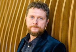 Taras Lyutyy, a man with reddish brown hair and beard and wearing a blue shirt and blazer, is seen leaning against a background of a wall made of bowed wood.