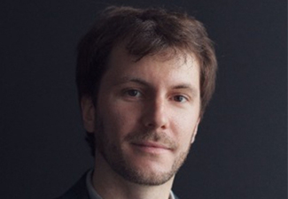 A young white man with a short bear and light brown hair, looking seriously into the camera wearing a button-down shirt and blazer