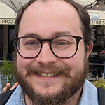 Close-up of a smiling Tom Rylett in front of a cafe. He has short, reddish brown hair with a receding hairline, a short beard, and is wearing glasses and a blue button-down shirt under a sweater.