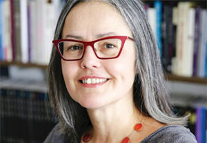 Close-up of Valerie Tiberius, a middle-aged white woman with long gray hair and red glasses, smiling and wearing a gray top and a bright red necklace standing in front of a bookshelf.