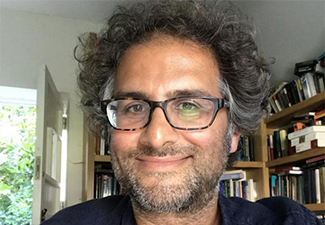 Headshot of Victor Tadros, bespectacled and with wild gray curls, smiling into the camera against a backdrop of a home library with a door opening to the green outside