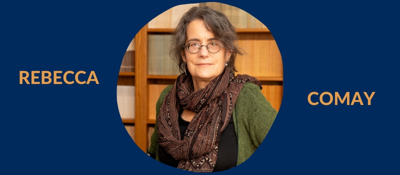 Head shot of Rebecca Comay, a middle-aged woman with medium-length, curly gray hair wearing glasses, a black top, green cardigan, and big patterned scarf, standing in front of a bookshelf. All this on a U of T blue background and her name in light orange.