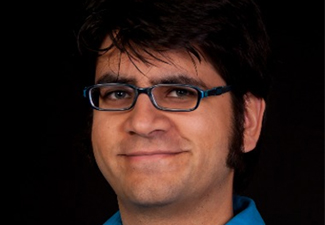 A close-up of a person with short black hair, sideburns, and glasses, smiling into the camera wearing a royal blue button-down shirt.