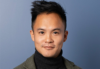 Head shot against a blue background of a young East Asian man wearing a black turtleneck under a tweed blazer.