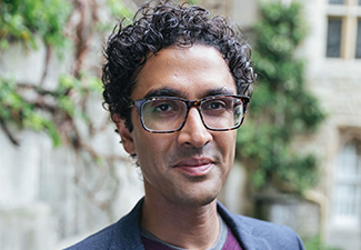 Head shot of a smiling Anil Gomes, a brown man with curly black hair and glasses wearing a sweater and tweed jacket standing in front of greened wall.