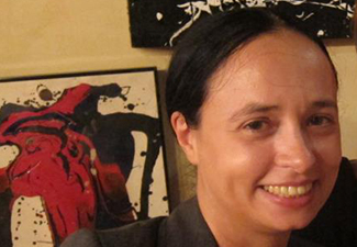 A smiling woman with straight brown hair made in a a bun, standing in a brown blazer in front of a wall of modern art.