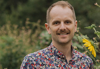 Head shot of David James Barnett, a white man with blond hair and mustache, standing smiling beside a sunflower wearing a casual flower-print button-down shirt