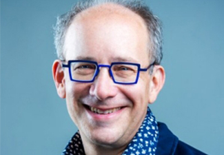 Close crop of David Morris, a middle-aged man, clean-shaven with receding gray hair, wearing bright blue spectacles, a blue flower-print shirt, and a blue blazer, smiling broadly into the camera.