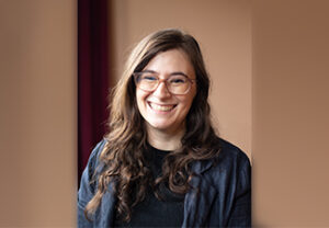Elena Gordon, smiling broadly and wearing glasses in front of an open, brownish background