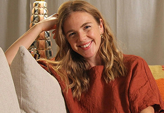 Head shot of Ellie Anderson, a young white woman with long blonde hair smiling brightly into the camera wearing a burnt-orange top while leaning against beige couch cushions.