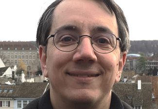 Close-up on a middle-aged man with short gray hair and glasses smiling into the camera against a backdrop of what looks like a small European town or city.