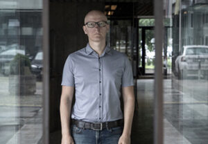 Gregor Moder, a bald white man with glasses dressed in short-sleeved shirt and jeans, stands in the middle of a glass doorway, showing urban reflections to both sides and the interior of the building in the middle. Tonally, everything is monochromatically blue-gray.