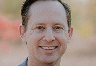 A middle-aged white man with short brown hair smiling into the camera, wearing a blue summer blazer