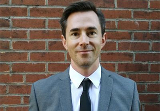 A young, clean-shaved white man with short, brown hair and wearing a business suits stands with a light smile in front of a brick wall.