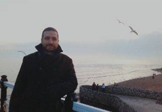 James Madaio, white, bearded man with short hair, leaning against an outdoor railing against a backdrop of sea and seagulls.