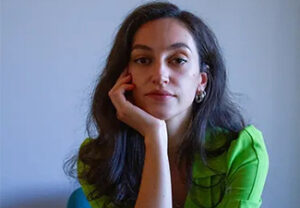 Head shot of Jeta Mulaj, a woman with long, thick black hair wearing a bright green shirt and looking seriously into the camera, her chin resting on on hand, her elbow propped up.