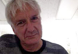 Screen shot of John Campbell, an older white man with short white hair wearing a gray T-shirt peering down at the camera.