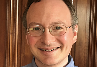 Close-up of a middle-aged man with short, graying dark hair and glasses, smiling into the camera wearing a blue button-down shirt against the backdrop of wood paneling.