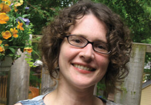 Head shot of Kara Richardson, a white woman with short, curly brown hair and glasses, wearing a summer dress and standing smiling in front of an outdoor garden wall with blooms.