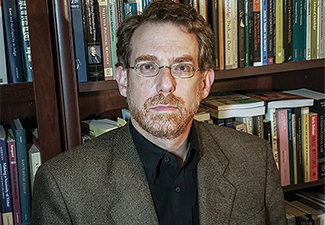 Head shot of a serious Lawrence Pasternack, a white man with reddish blond hair, short beard, and glasses, sitting in front of a bookshelf wearing a black shirt and brown blazer.