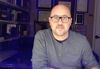 Screen shot of Mark Schroeder, a bald white man with glasses and a beard, sitting in a home office wearing a gray sweater.