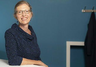 A photo of Pauline Kleingeld, an older white woman with blonde hair and glasses, sitting smiling at a desk, with a blue wall and a coatrack visible behind her