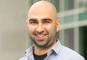 Head shot of a smiling Reza Hadisi sporting a light beard and wearing a black T-shirt topped by a light blue shirt