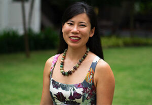 Head shot of Robin Zheng, an East Asian woman with long black hair wearing a colorful summer dress and necklace of large beads standing on a lawn and smiling.
