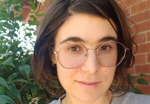 Close crop of Sara Aronowitz, who has medium-length borwn hair and glasses, standing against an outside brick wall wearing a gray T-shirt.