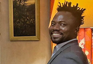 A smiling black man with short dreads and a goatee stands in a warmly lit interior wearing a suit and turning toward the camera.