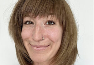 Close-up of a person with shoulder-length straight brown hair, dark brown eyes, and a nose ring, smiling into the camera wearing a dark, V-neck top and delicate cold chain.